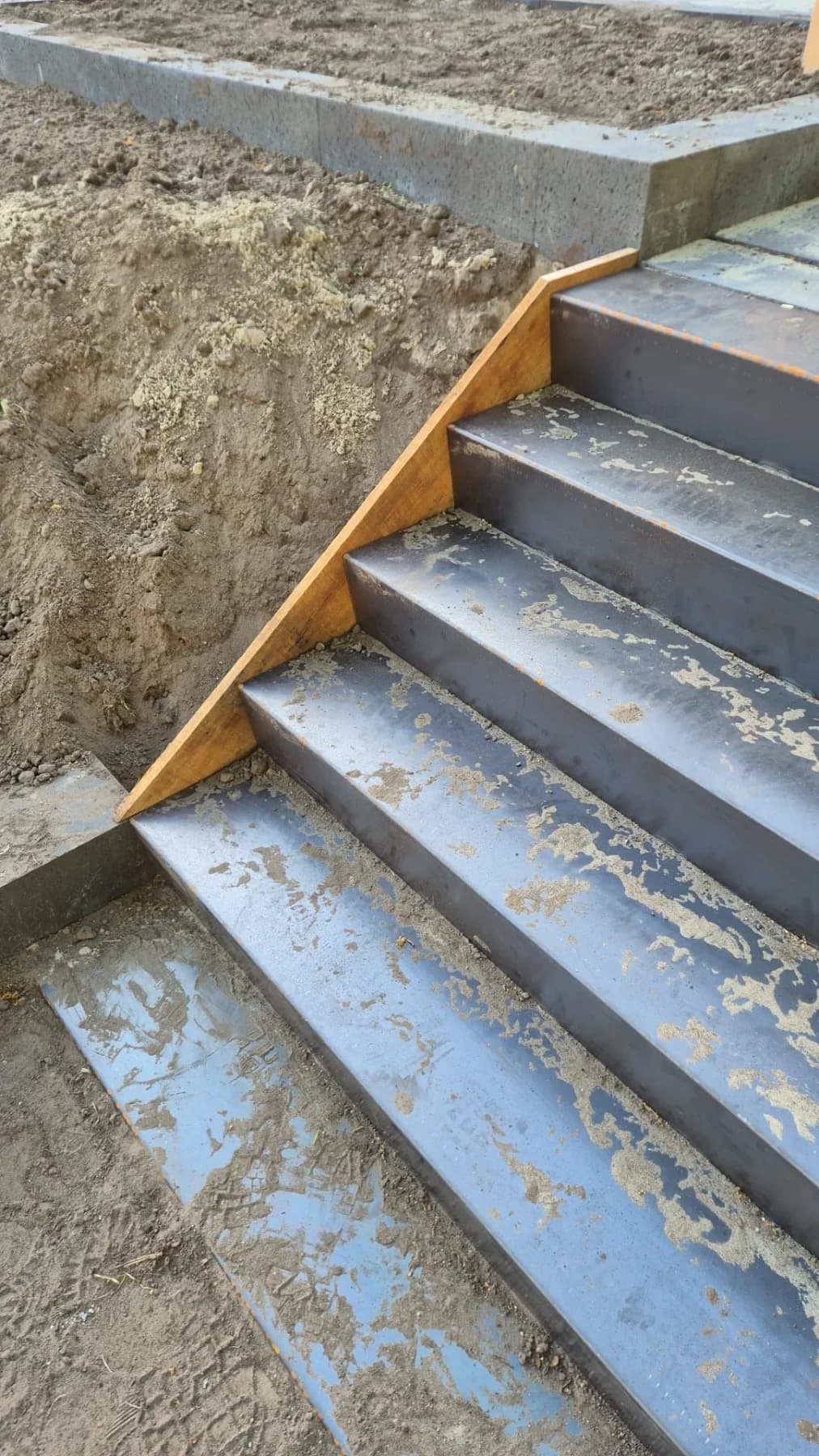 Stalen treden van de nieuwe trap van dichtbij, met zand erop en een oranjebruine plank langs de zijkant, tijdens de aanleg in Oldeberkoop