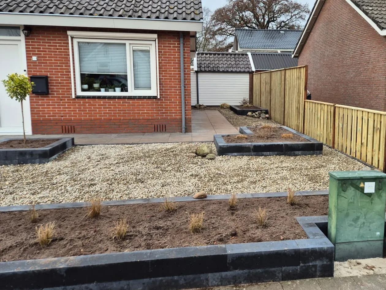 Onderhoudsarme voortuin met grind in Harkema van dichtbij: verhoogde bakken van stapelblokken met jonge siergrassen langs de stoep en een tegelpad langs de gevel van de vrijstaande woning