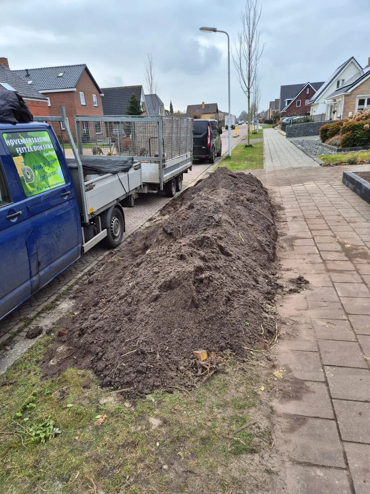 Bus van Hoveniersbedrijf Feitze Dijkstra met aanhanger naast een lange hoop afgegraven grond langs de stoep in Harkema