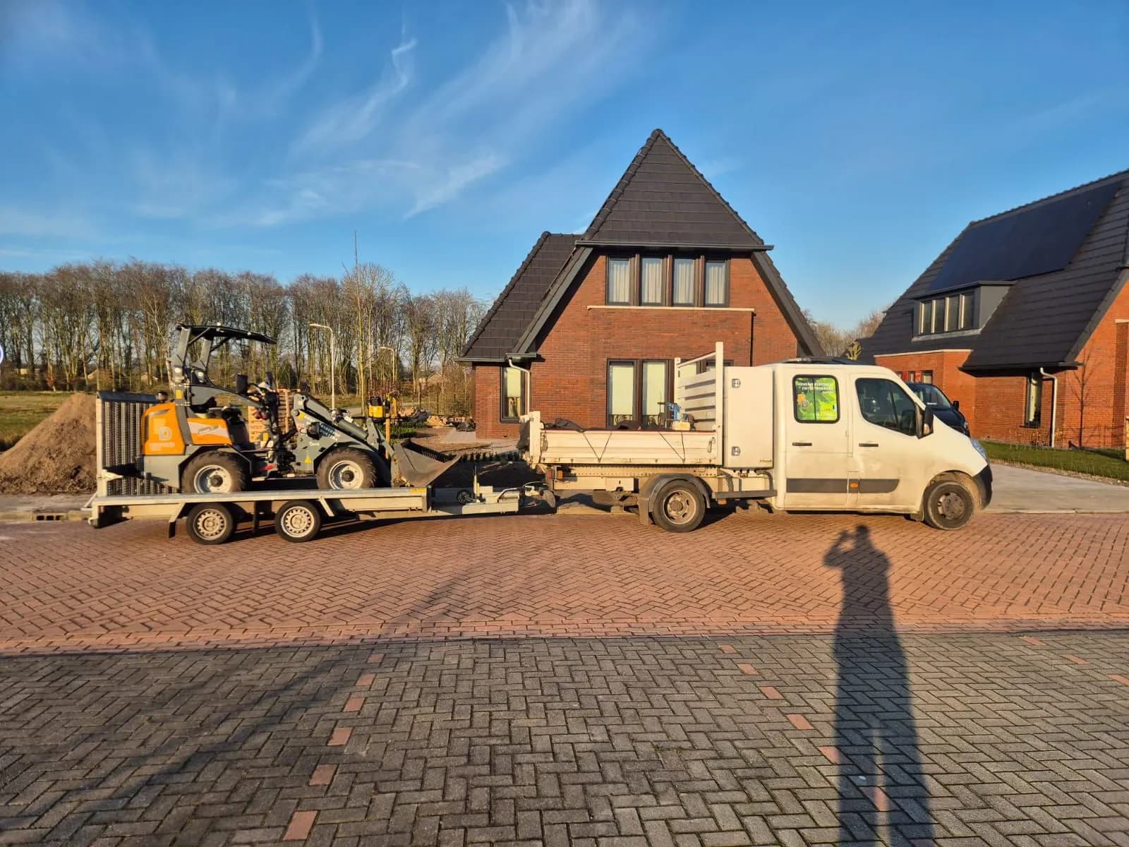 Shovel op een aanhanger achter een bus in een nieuwbouwwijk in Gorredijk, met een hoop zand ernaast, bij de start van de aanleg van de Japanse tuin