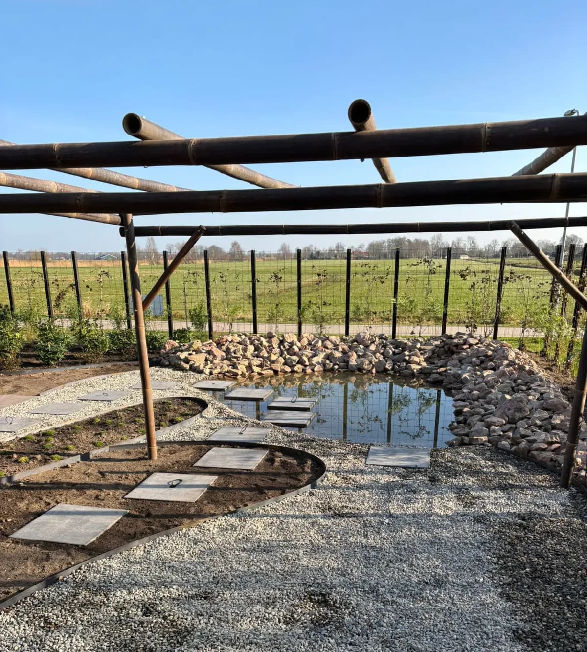 Japanse tuin met een vijver omringd door rotsen, een grindpad met betontegels en een pergola van bamboe, met uitzicht op het Friese landschap aan de rand van Gorredijk