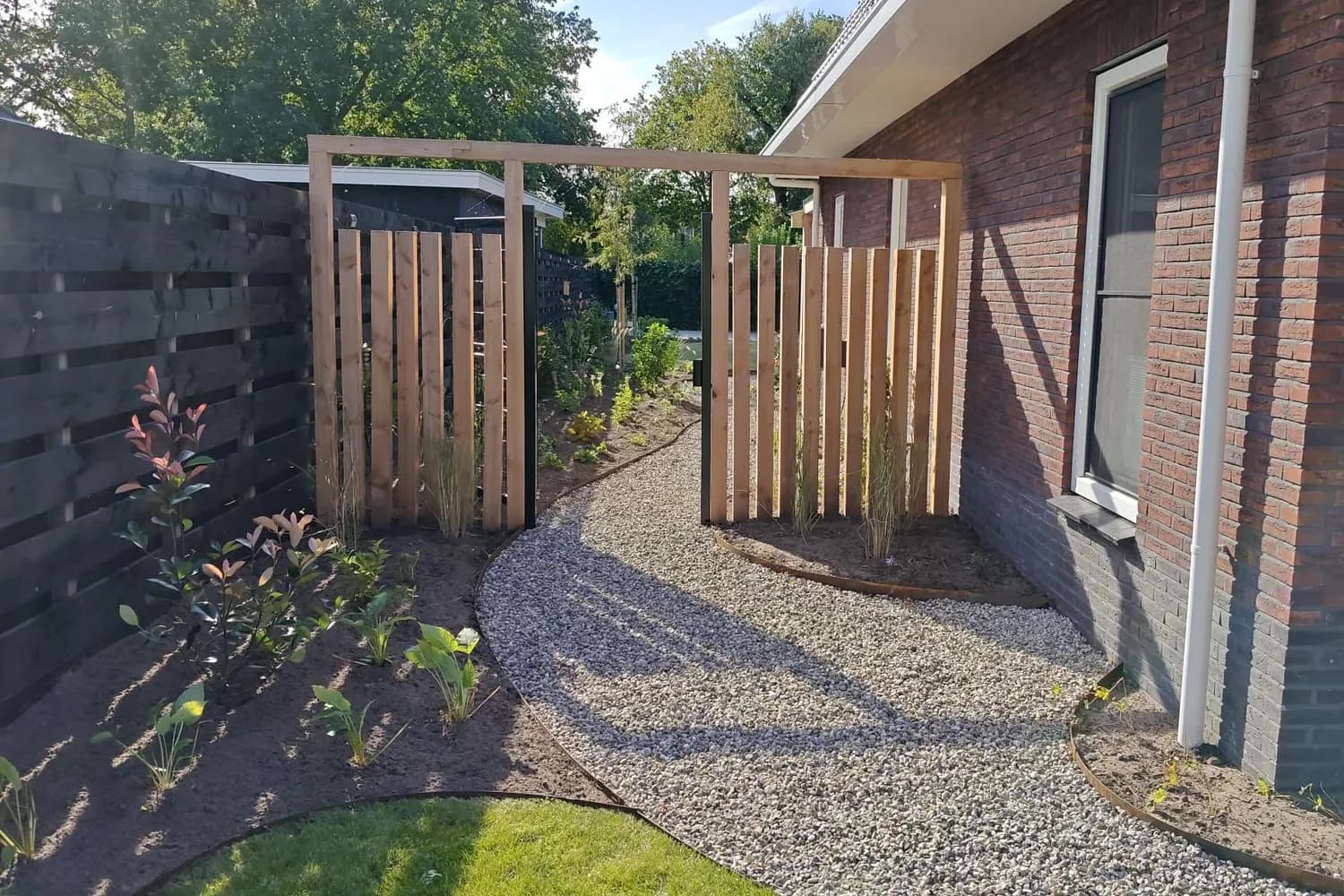 Grindpad met rand van cortenstaal langs het huis in Lippenhuizen, met een doorgang onder een pergola van douglas tussen twee houten schermen