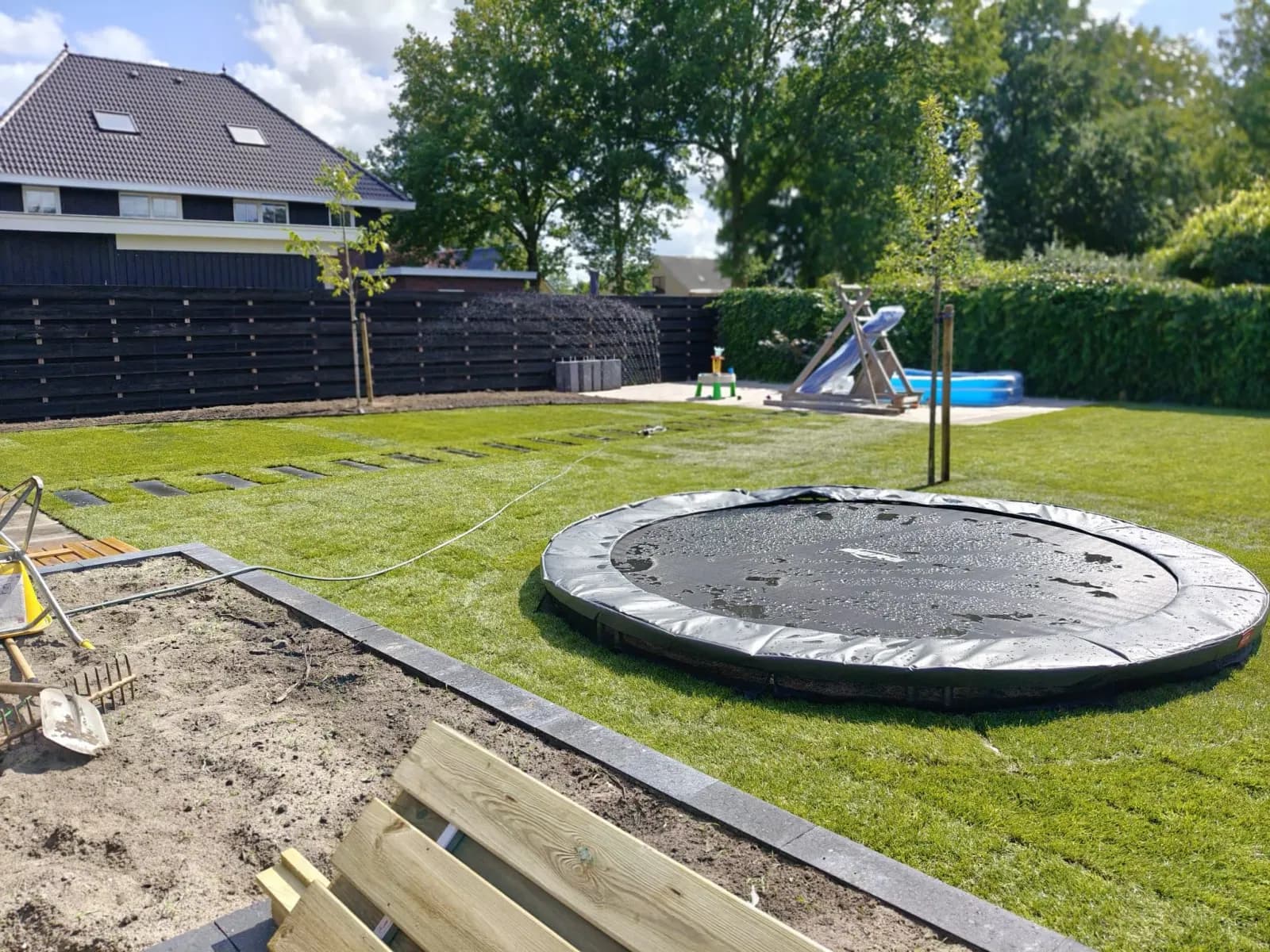 Pas gelegde graszoden met zichtbare naden rond een trampoline in de grond in de familietuin in Lippenhuizen, met een zandbak op de voorgrond