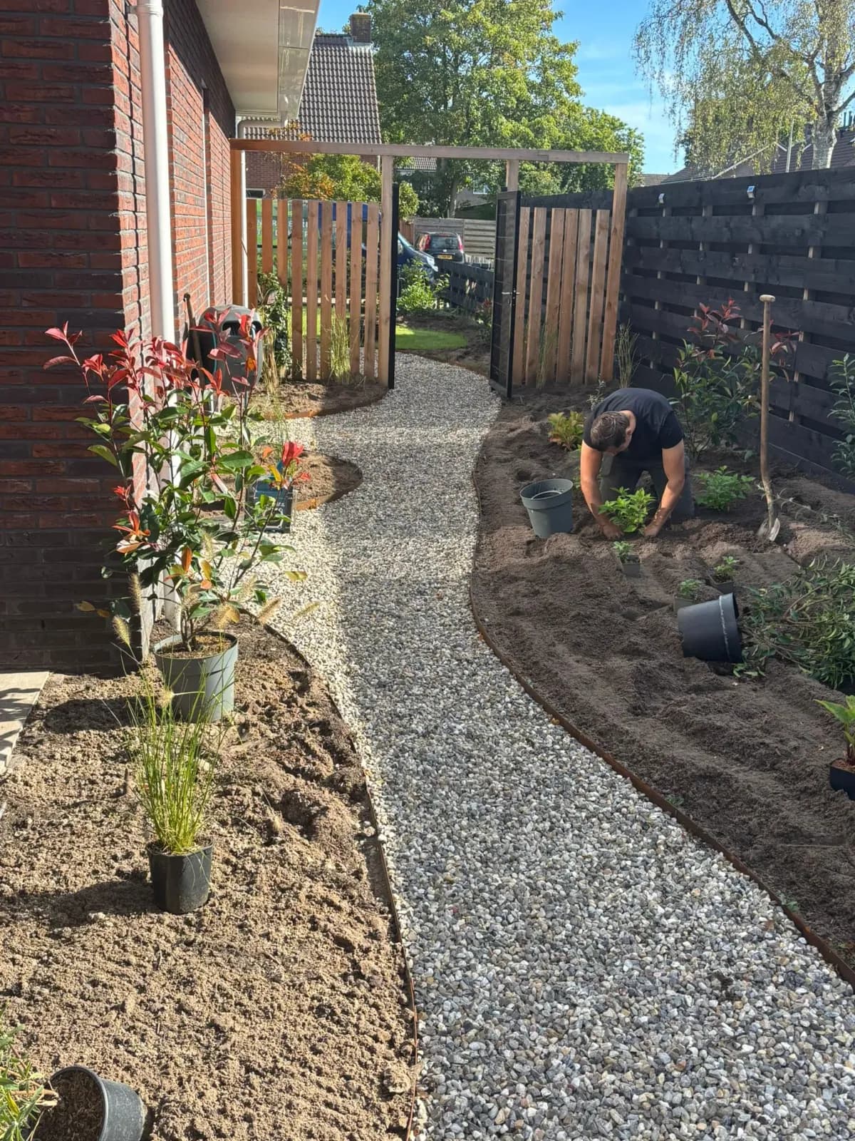 Grindpad langs de gevel naar de houten poort in Lippenhuizen, met jonge heesters in de nieuwe plantvakken en achterin een hovenier aan het planten