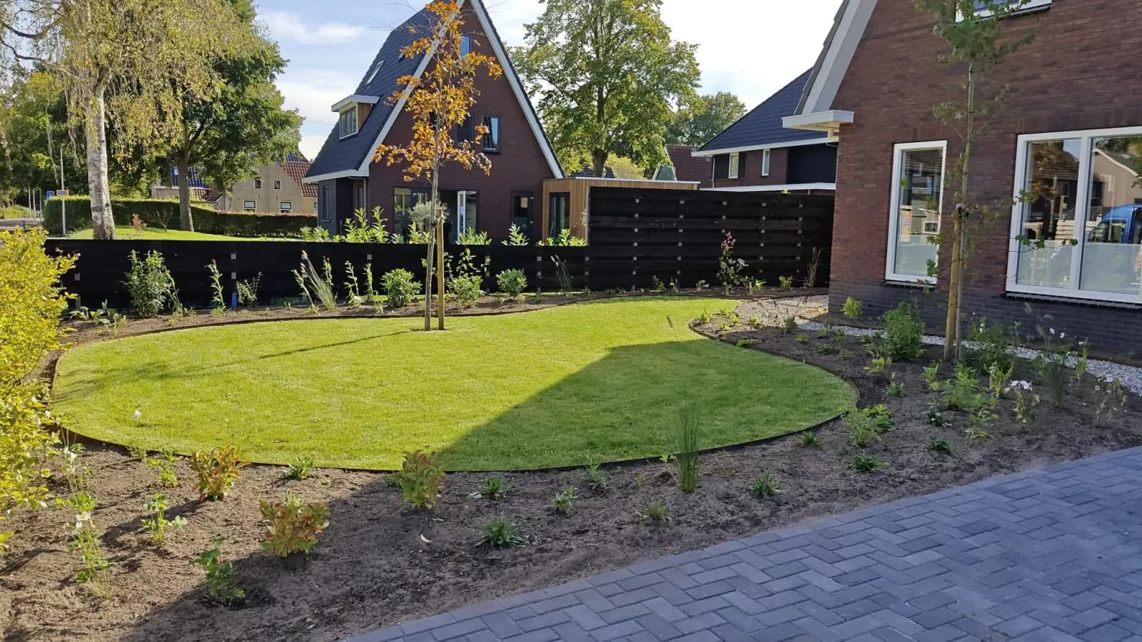 Nieuw aangelegde voortuin met een rond gazon, een jonge boom en net geplante borders bij een vrijstaande nieuwbouwwoning in Lippenhuizen