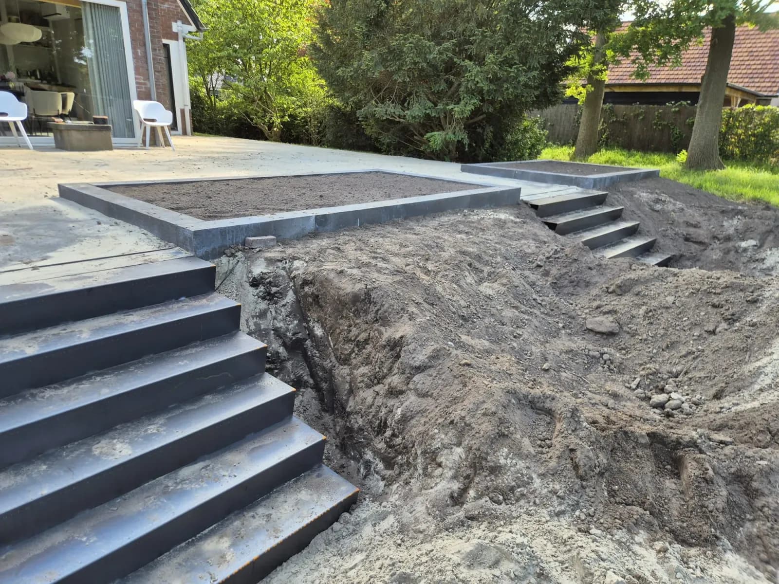 Vers uitgegraven grond naast de nieuwe trap tijdens het grondwerk in de tuin in Oldeberkoop, met een verhoogd plantvak langs het terras en verderop een tweede trap