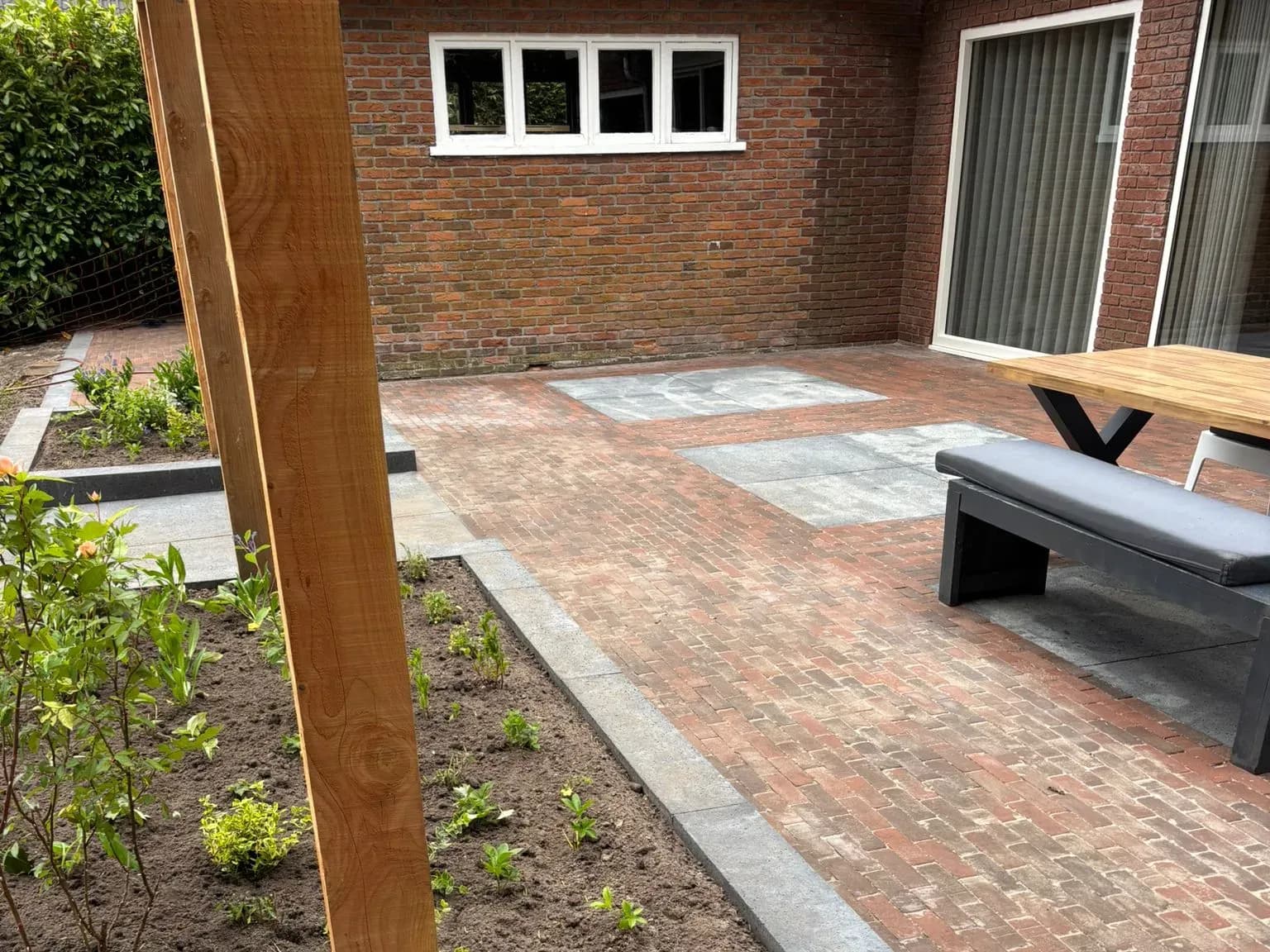 Terras in Oldeberkoop na oplevering: gebakken klinkers met vakken van oud-Hollandse tegels, een bank en tafel, een antraciet band langs een jonge border en de houten palen van de pergola