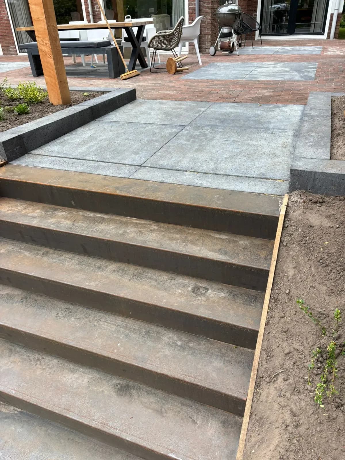 Trap van het terras naar de lagere tuin in Oldeberkoop, van bovenaf: cortenstalen treden, een bordes van grote tegels en daarachter het terras van gebakken klinkers met tafel en barbecue