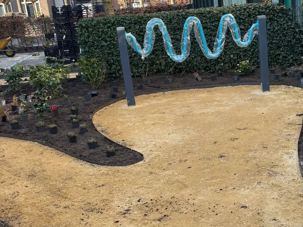 Breed slingerend pad van gele halfverharding met een stalen rand, jonge planten in potjes in de border en een blauwe spiraal tussen twee donkere palen, tijdens de aanleg van de belevingstuin bij zorglocatie Voltawerk in Gorredijk