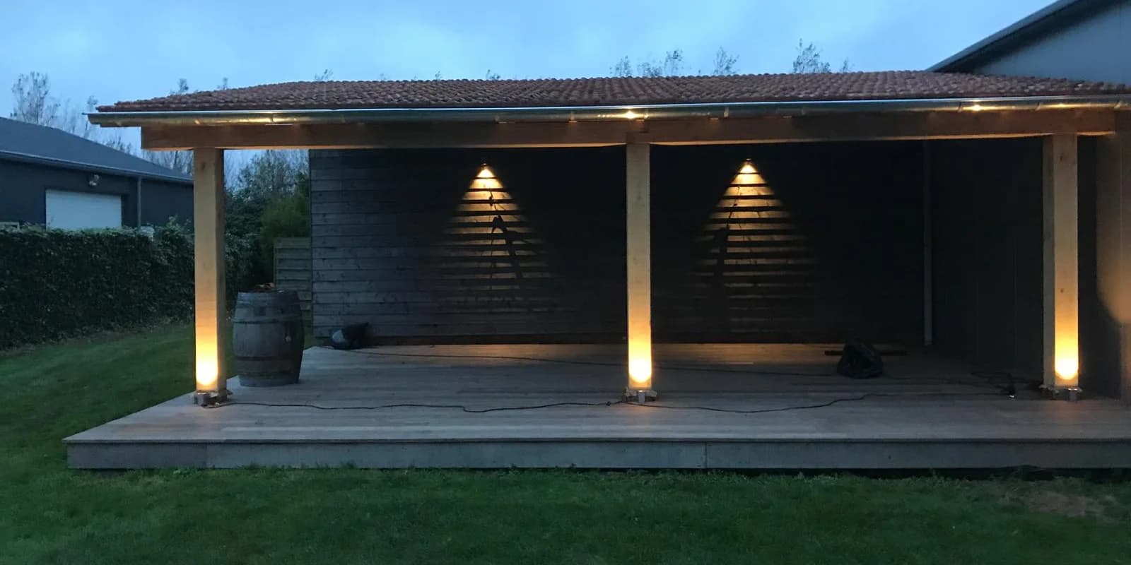 Veranda met een pannendak en een donkere achterwand in de schemering. Onder aan de staanders en op de wand branden lampen, links staat een houten ton