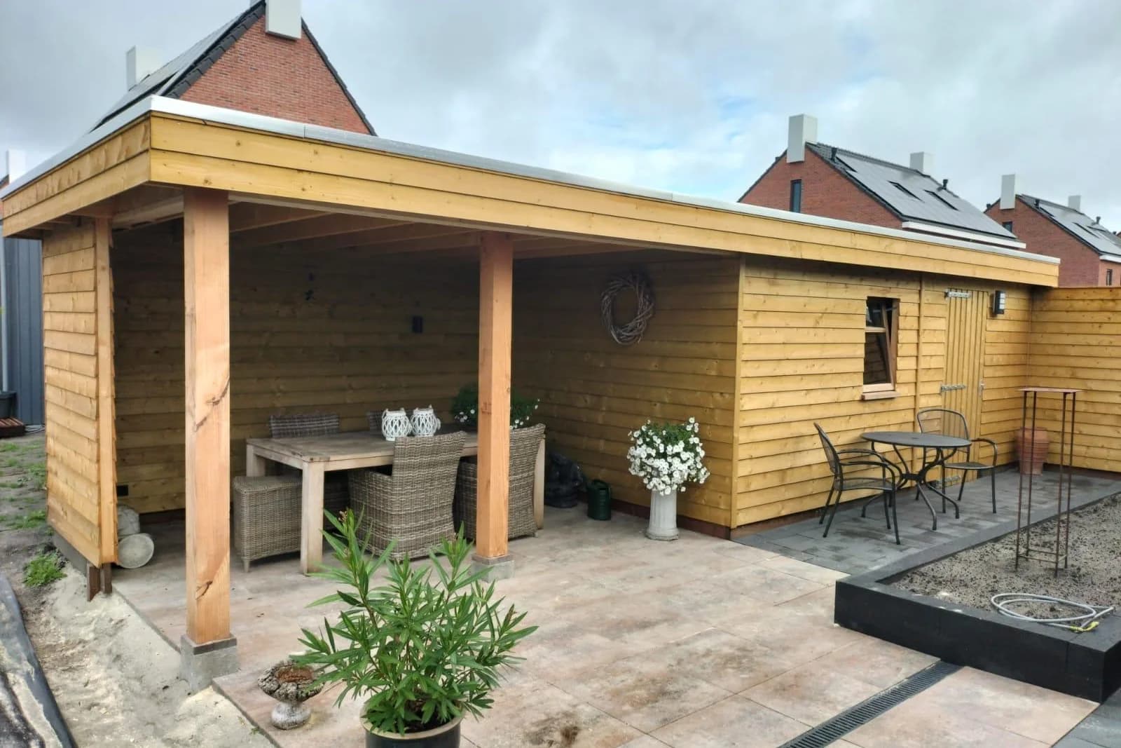 Houten overkapping met een dichte achter- en zijwand en een tafel met rieten stoelen eronder. Onder hetzelfde dak zit een berging met een raam en een deur, ervoor ligt een terras van grote tegels
