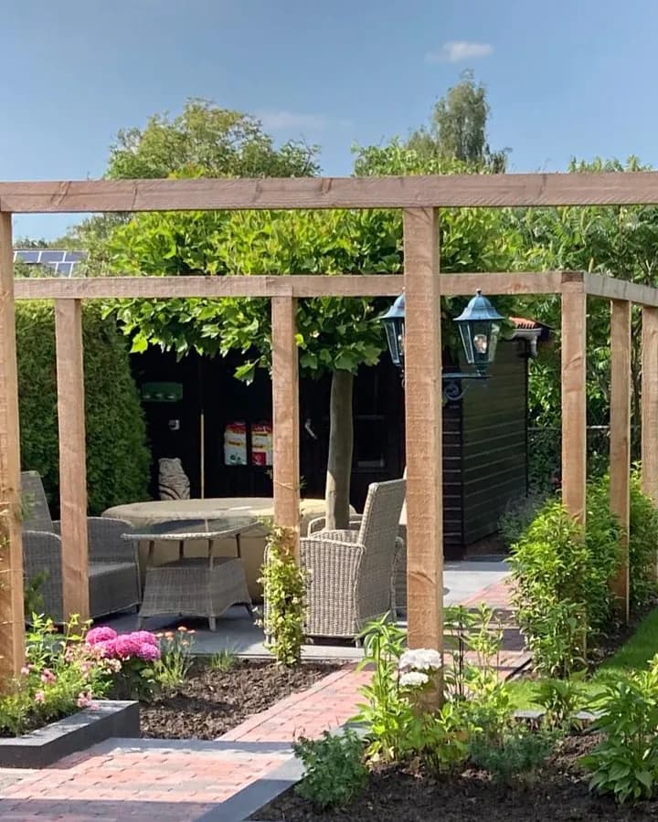 Pergola van douglas boven een zithoek met rieten stoelen en twee lantaarns, met daarachter een zwart bijgebouw, ervoor borders in een donkere rand en een pad van gebakken klinkers