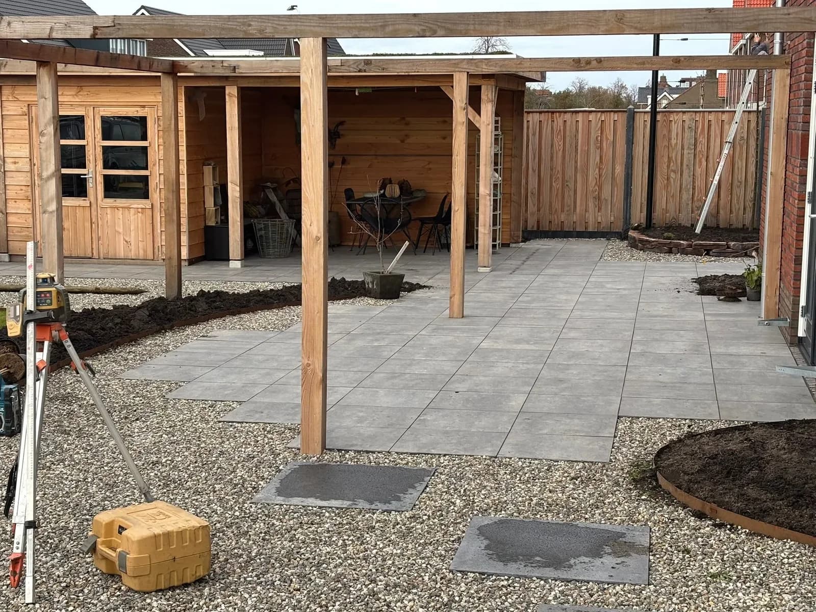 Nieuwbouwtuin in Gorredijk tijdens de aanleg: een houten overkapping met berging achter een terras van grijze tegels, ervoor een pergola, grind met staptegels en een meetlaser op een statief