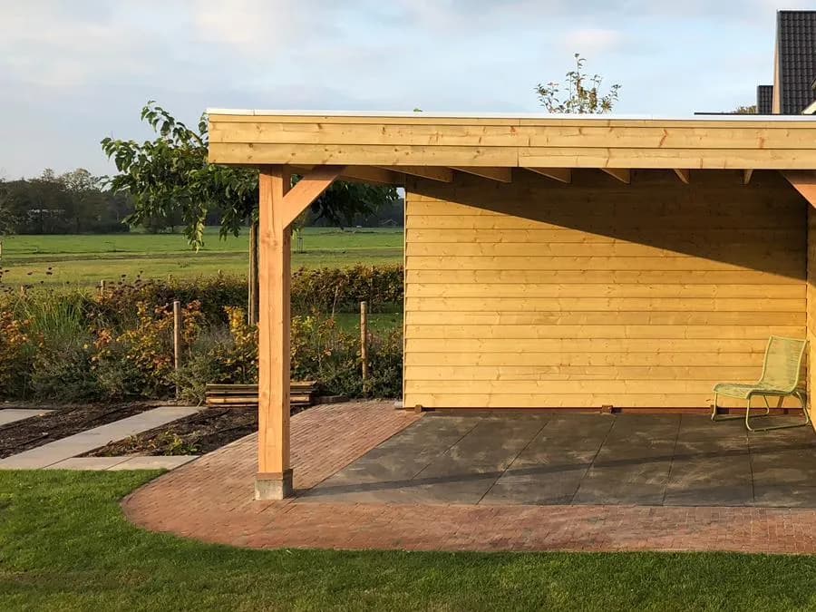 Vrijstaande houten overkapping met een schoor aan de staander en een stoel onder het dak, met een pad van gebakken klinkers, gazon en een border voor het weiland, in het late middaglicht