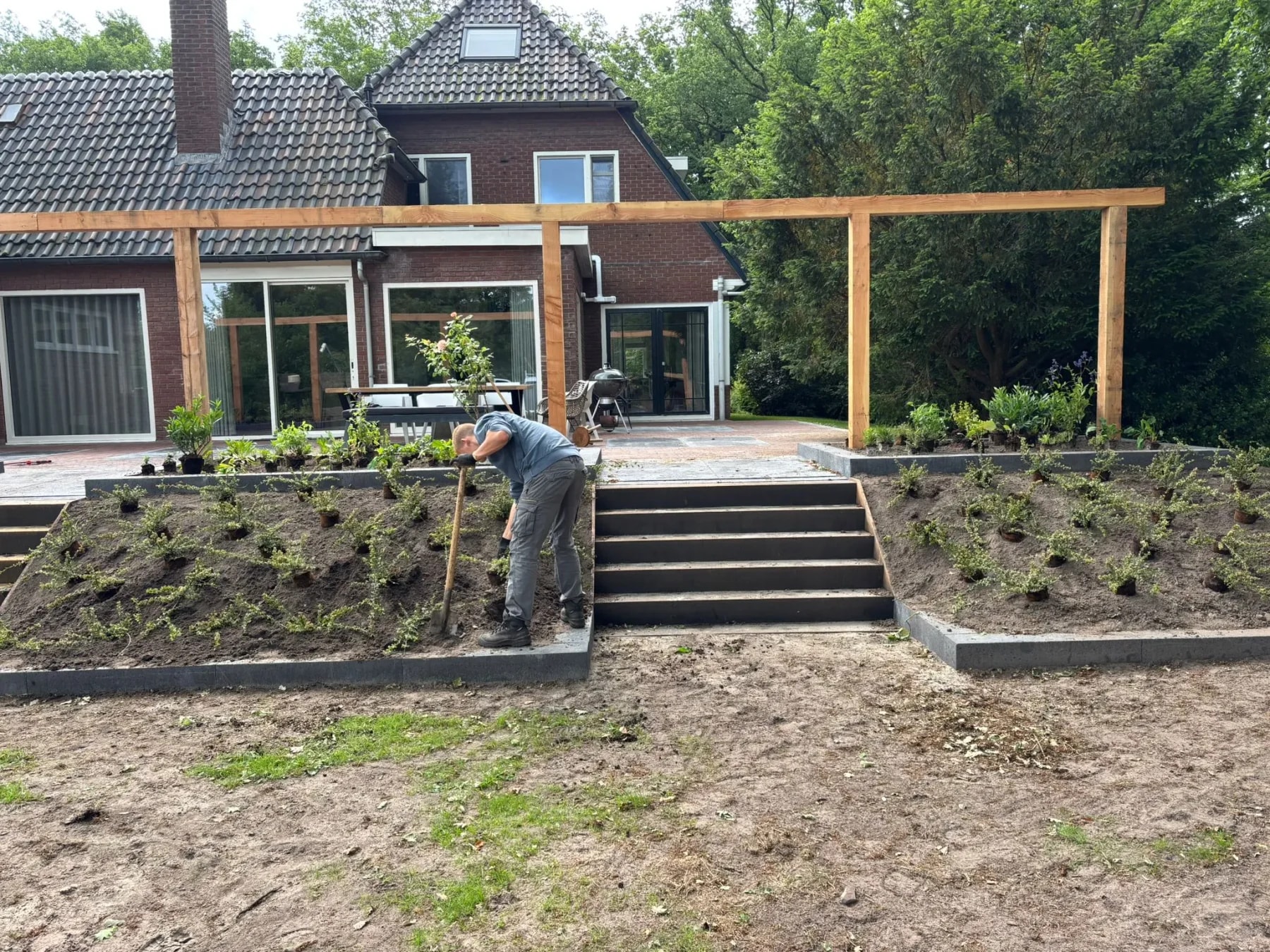 Terras met een trap naar de lagere tuin en een pergola van douglas bij een woning in Oldeberkoop, gezien vanuit de tuin, met een hovenier die jonge struiken plant op de schuine border naast de trap