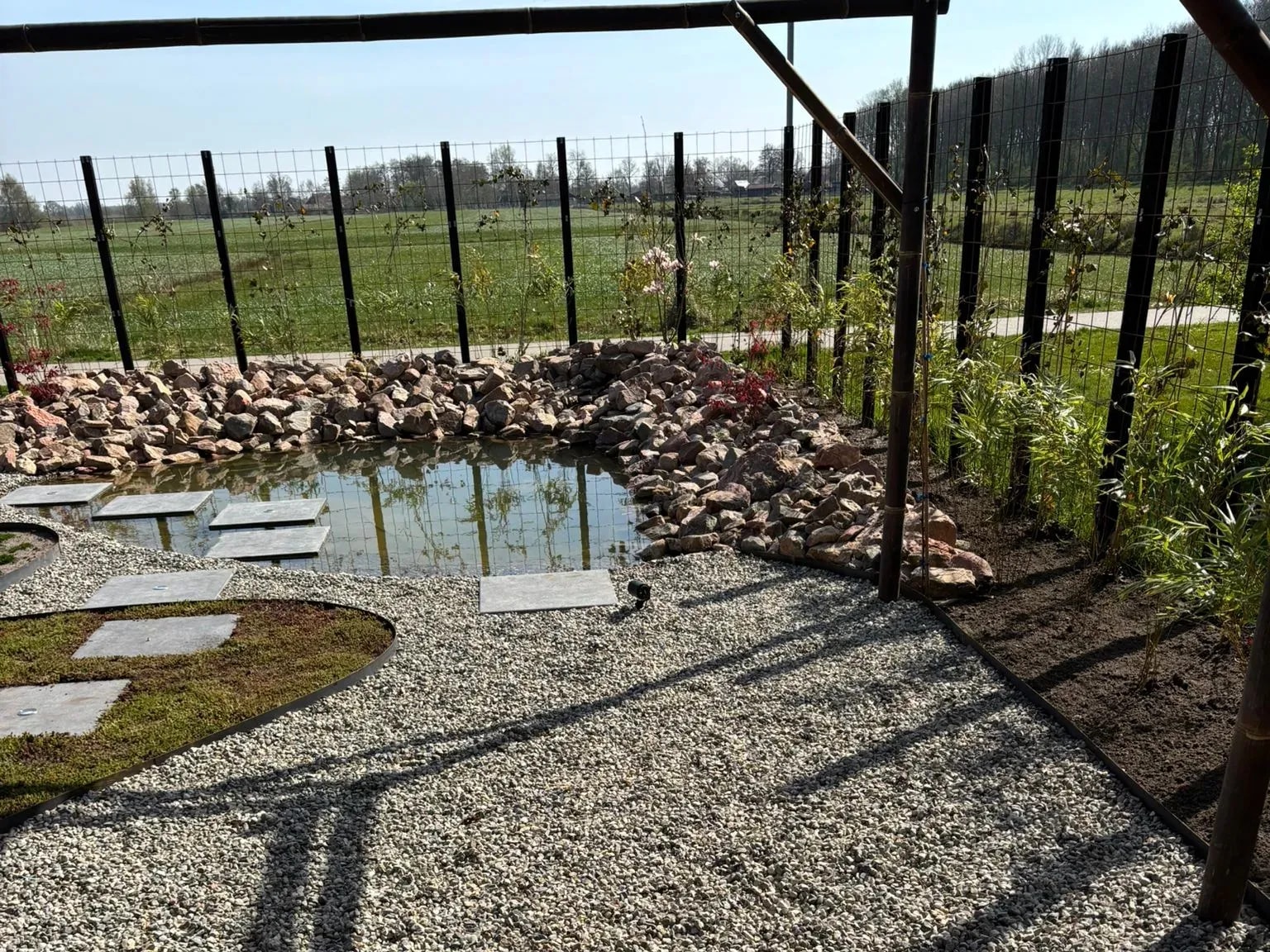 Vanaf het terras onder de pergola van bamboe: grind, staptegels en de vijver met rotsrand in een Japanse tuin bij een nieuwbouwwoning in Gorredijk, met het open weiland erachter