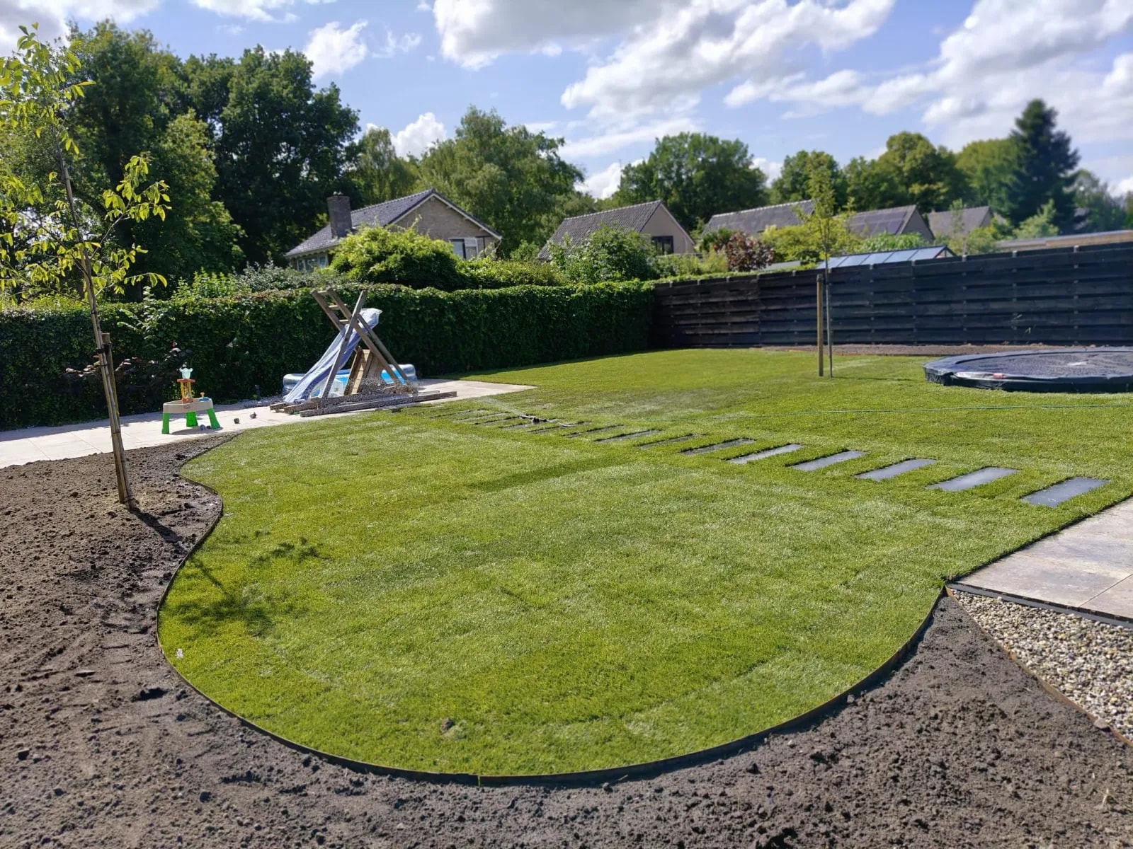 Kindvriendelijke nieuwbouwtuin in Lippenhuizen met een gebogen gazon, rechte staptegels, een speeltoestel en een trampoline in de grond, omringd door een haag en een zwarte schutting