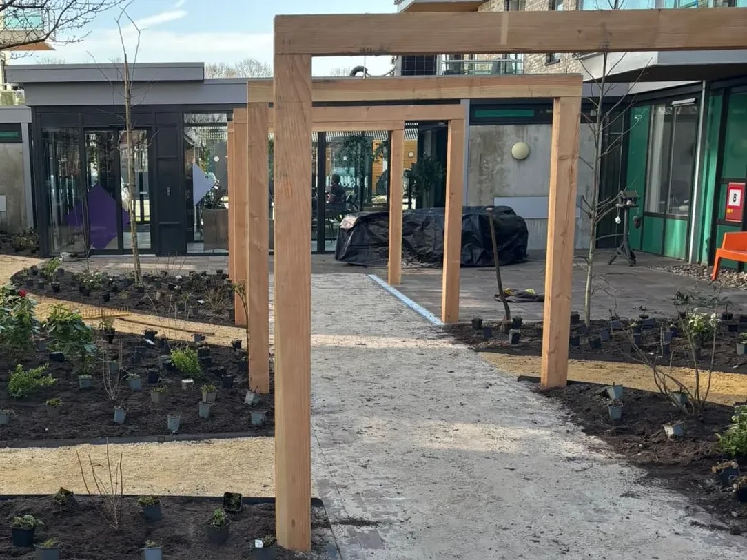 Houten pergola over de hoofdroute door de nieuw aangeplante belevingstuin bij zorglocatie Voltawerk in Gorredijk, met kale bomen tussen de gebouwen