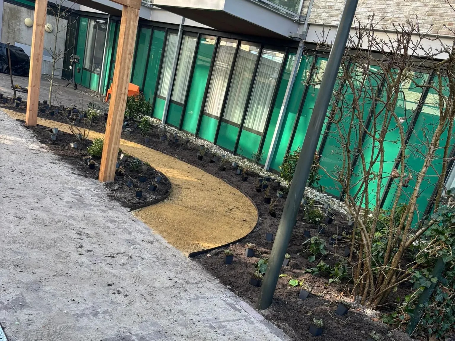 Slingerend pad van gele halfverharding met een houten pergola en nieuw aangeplante borders in de belevingstuin bij zorglocatie Voltawerk in Gorredijk