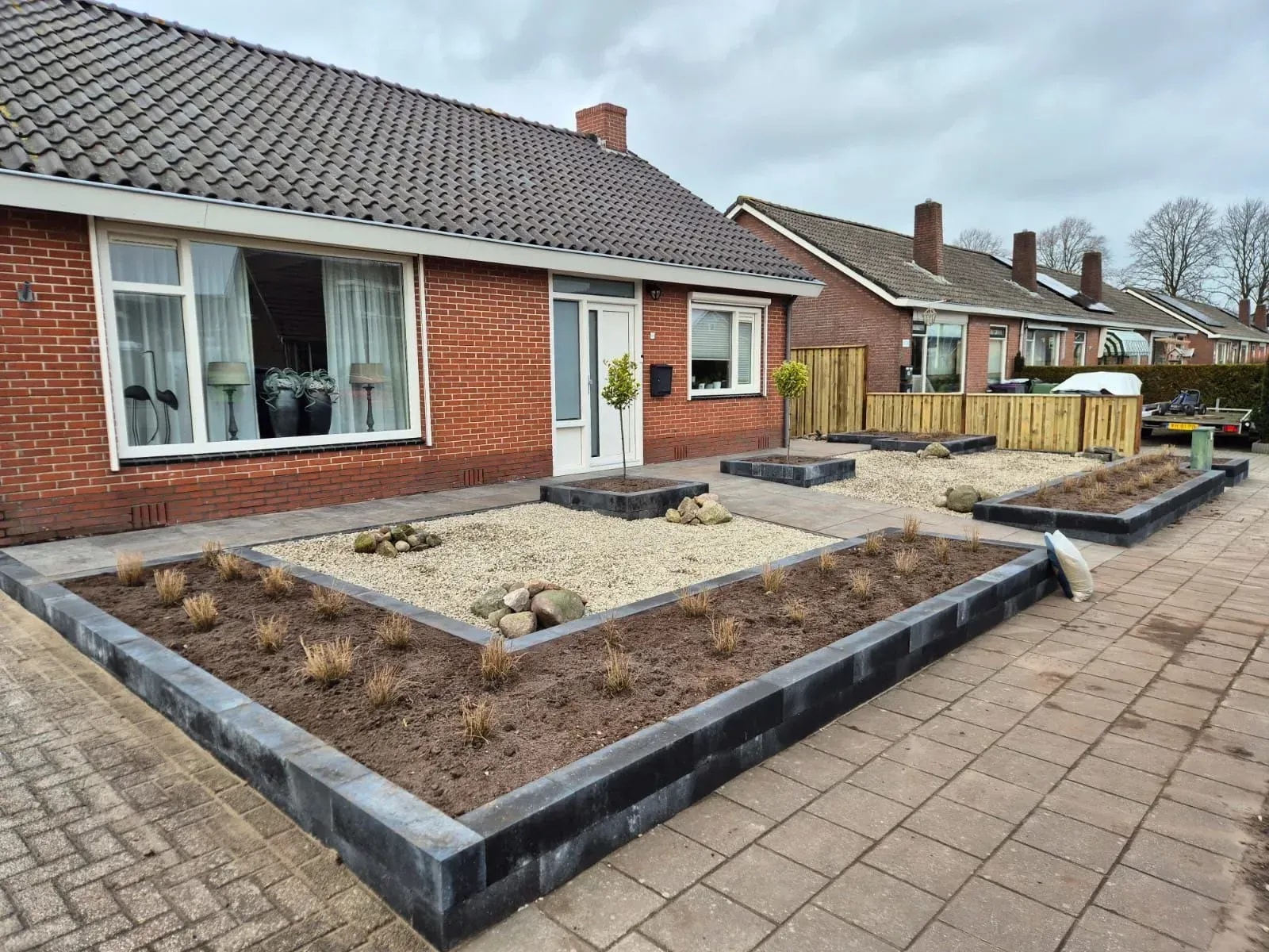 Onderhoudsarme voortuin met grind en verhoogde bakken van stapelblokken, met siergrassen en twee bolboompjes, bij een vrijstaande woning in Harkema