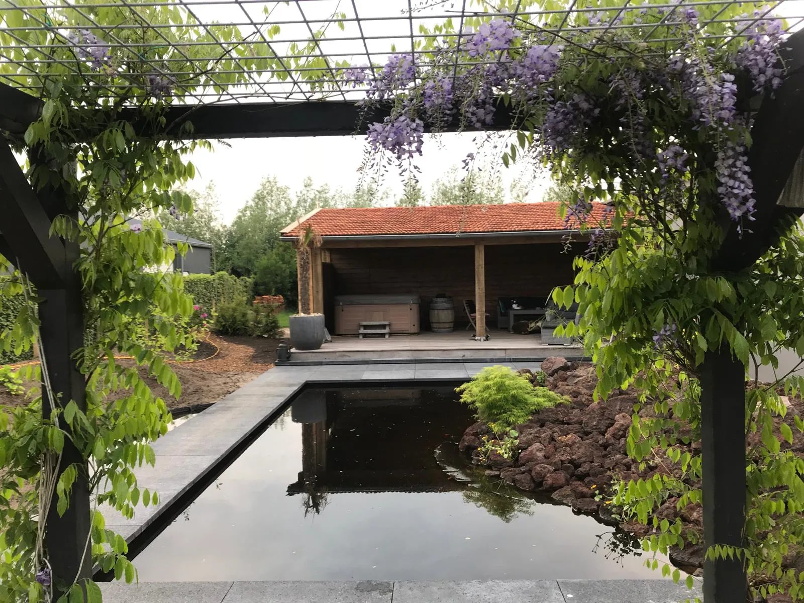 Veranda met een pannendak aan de overkant van een strakke vijver, gezien door een pergola met bloeiende blauwe regen