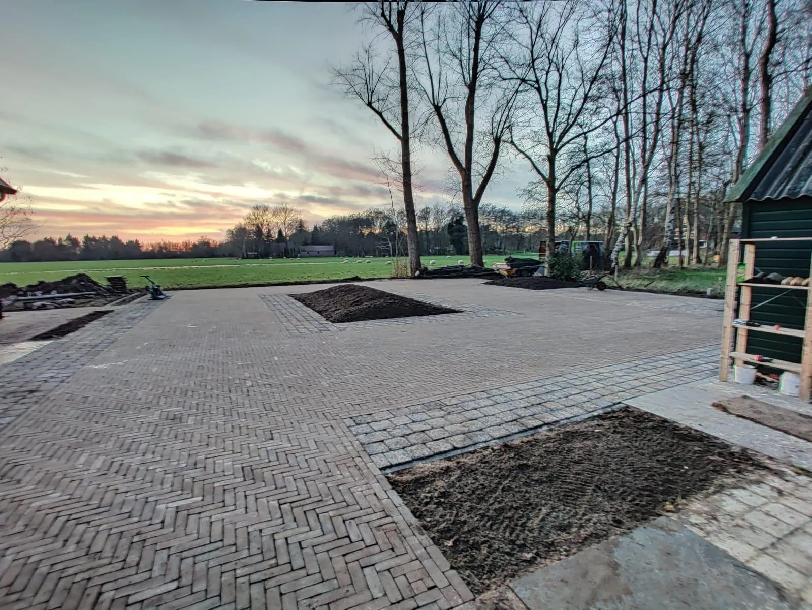 Erf van klinkers in visgraatverband met een plantvak met verse aarde, aan de rand van een weiland bij zonsondergang
