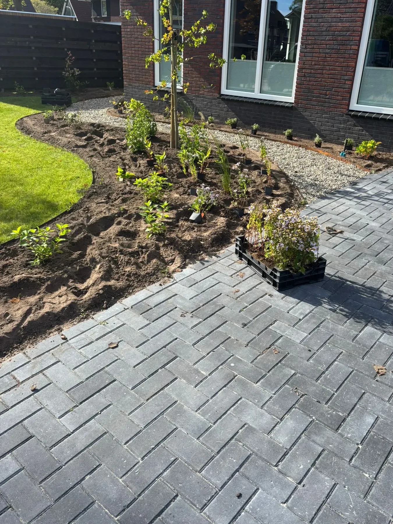 Oprit van antraciet klinkers in keperverband bij een woning in Lippenhuizen, met een pas aangeplante border en een krat planten op de stenen