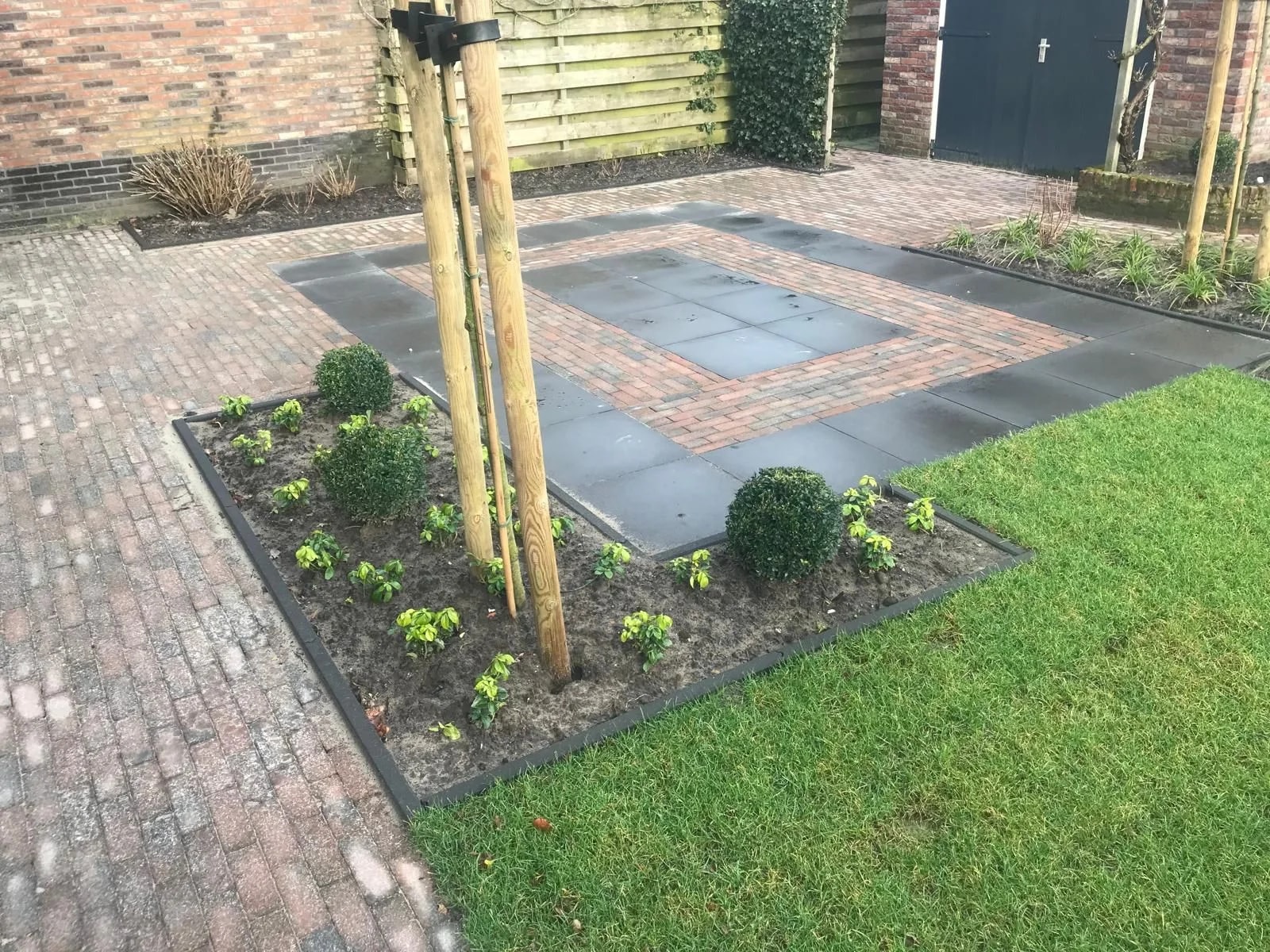 Terras van antraciet tegels met een bies van klinkers erin, tussen een border met buxusbollen en het gazon