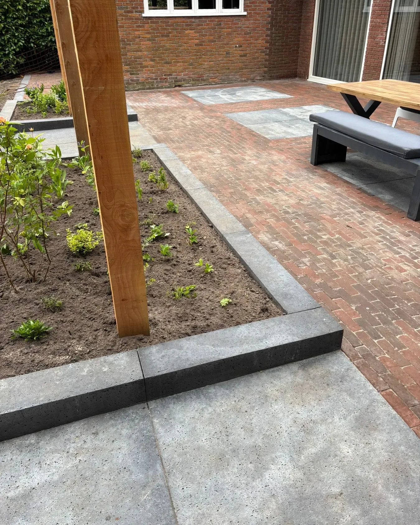 Terras in Oldeberkoop na oplevering: gebakken klinkers met vakken van grote grijze tegels, een antraciet band langs een jonge border en de houten paal van een pergola
