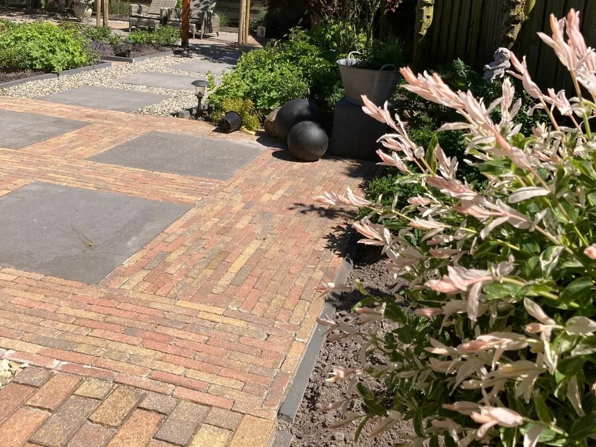 Zonnig terras van klinkers in rood en geel met vakken van grote grijze tegels, tussen borders met een rode esdoorn en leilinden