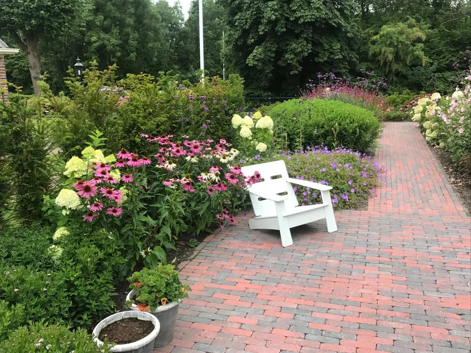 Pad van rode en grijze klinkers langs een border met hortensia, zonnehoed en ooievaarsbek, met een witte tuinstoel op de stenen