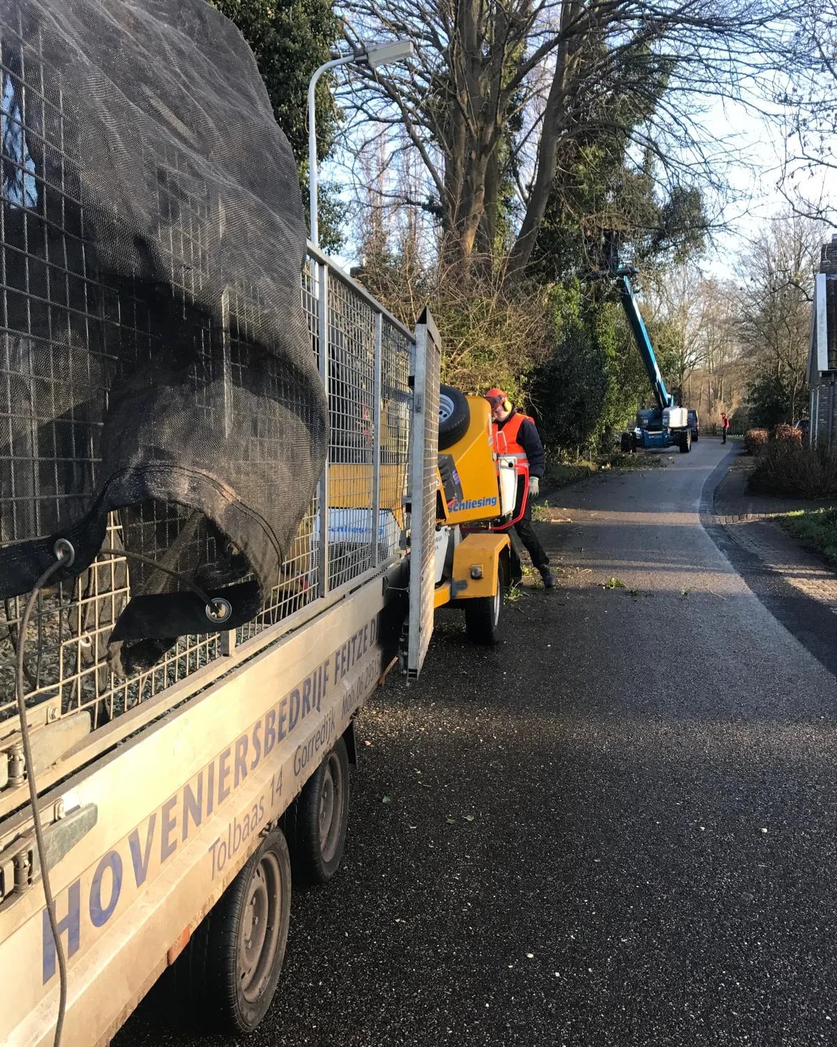 Aanhanger met het opschrift Hoveniersbedrijf Feitze Dijkstra langs een laan, met daarachter een versnipperaar en een medewerker met helm, gehoorkap en oranje hesje. Verderop werkt een hoogwerker in de bomen