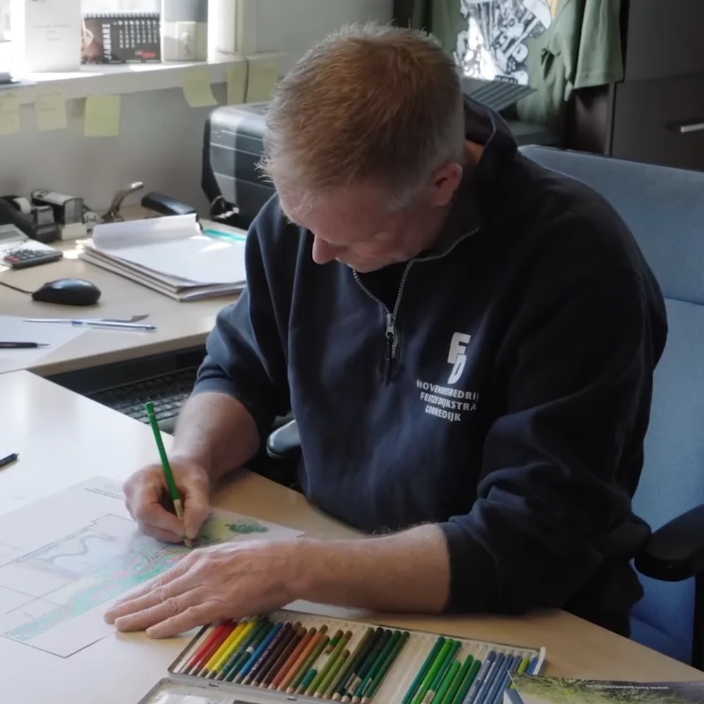 Feitze Dijkstra kleurt aan zijn bureau een tuinontwerp in met een groen potlood, in een trui met het logo van zijn hoveniersbedrijf