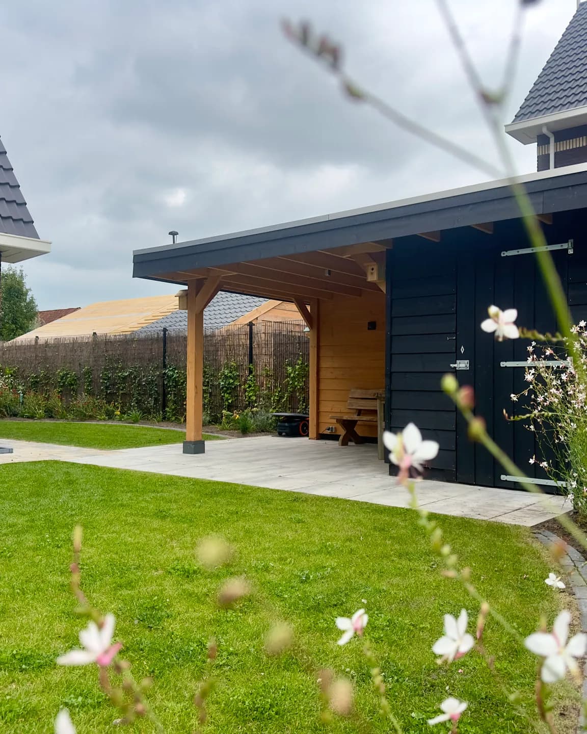 Achtertuin in Gorredijk met gazon, een terras van lichte tegels onder een houten overkapping met zwarte berging en witte gaura op de voorgrond
