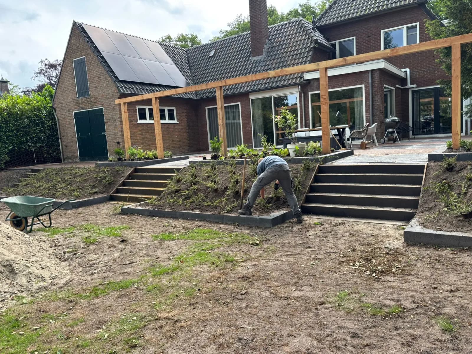 Hovenier aan het werk tussen twee trappen in een nieuw aangelegd talud achter een woonhuis met een houten pergola in Oldeberkoop