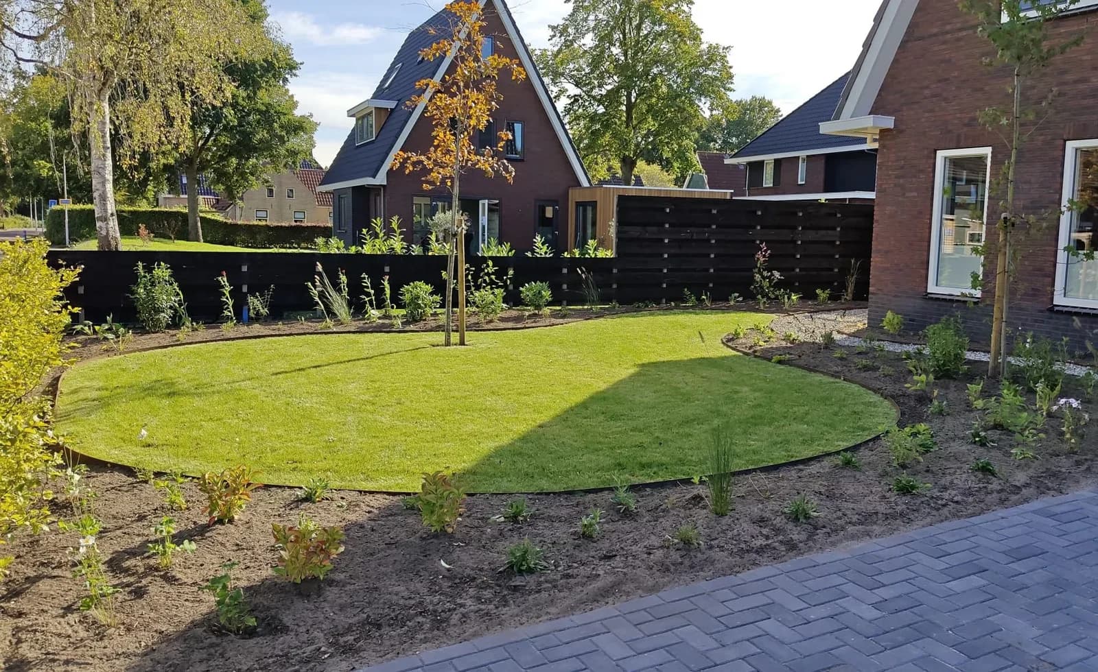 Tuin in Lippenhuizen met een rond gazon in een stalen rand, een jonge boom, borders met jonge planten langs een zwarte schutting en bestrating van donkere klinkers in keperverband
