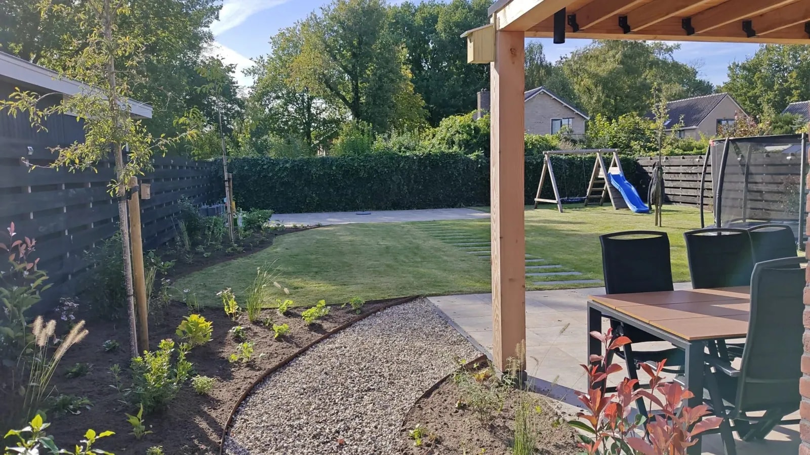 Familietuin in Lippenhuizen, gezien vanaf het terras onder de houten veranda: een speelgazon met staptegels, een schommel met glijbaan, een gebogen grindpad en nieuwe borders langs een zwarte schutting