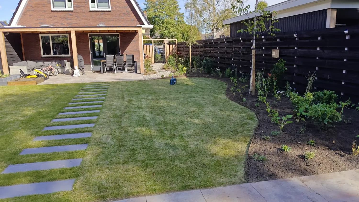 Achtertuin in Lippenhuizen met gazon, staptegels en gebogen borders langs een zwarte schutting