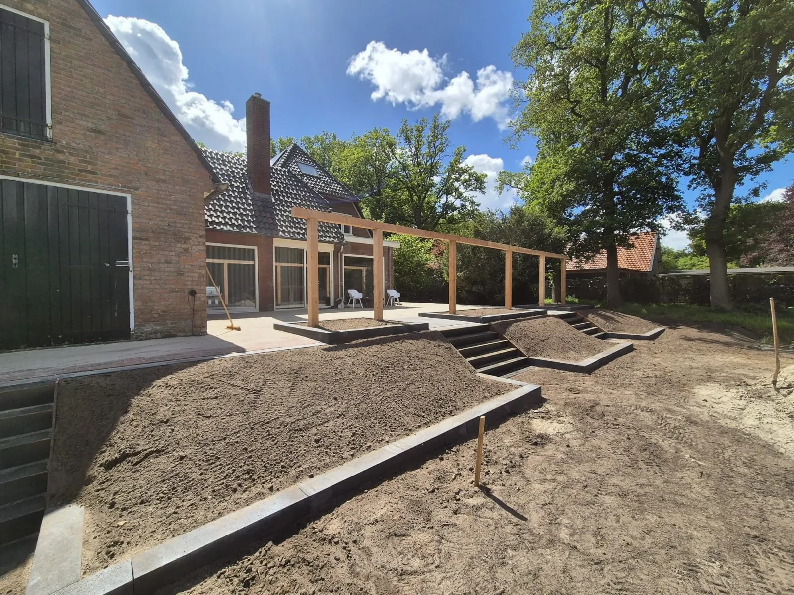Dezelfde tuin tijdens de aanleg: kale schuine borders in randen van grijze blokken, met trappen naar het verhoogde terras en de pergola. Daarachter het huis en hoge bomen.