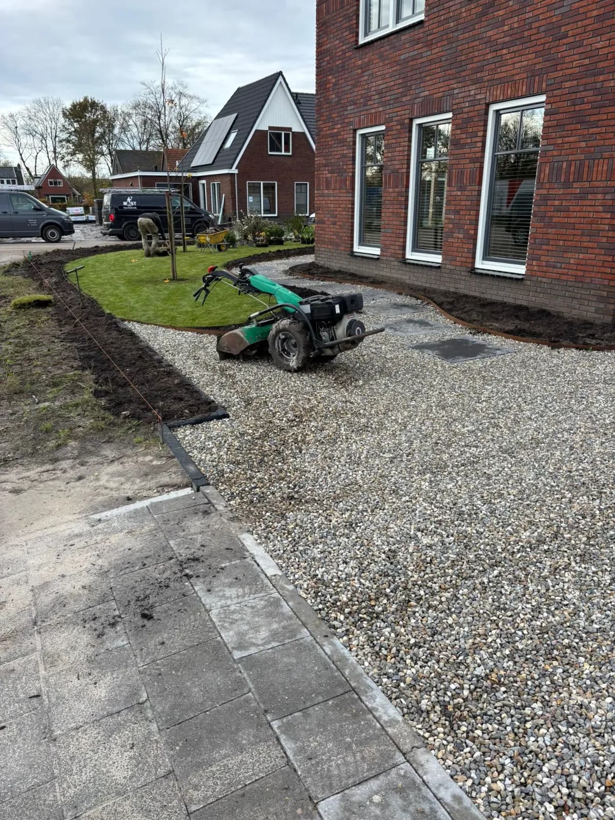 Grindpad met grote tegels langs de bakstenen gevel van een nieuwbouwwoning in De Vrieswijck in Gorredijk: een brede grindstrook tussen huis en gazon, omlijst door gras en verse aarde, met een tuinfrees en achterin een hovenier aan het werk