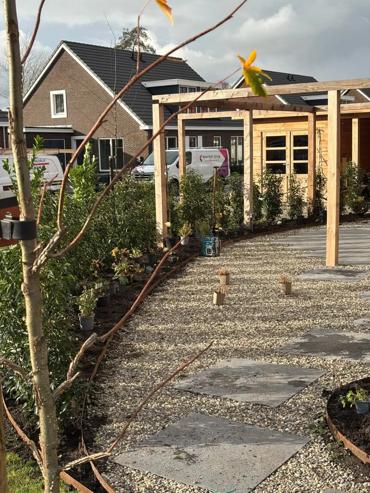 Gebogen grindpad met grote grijze staptegels langs een houten pergola, tijdens de aanleg. Links een plantvak met een rand van cortenstaal, waar de planten nog in pot klaarstaan. Op de achtergrond de houten schuur en nieuwbouwwoningen in de wijk.