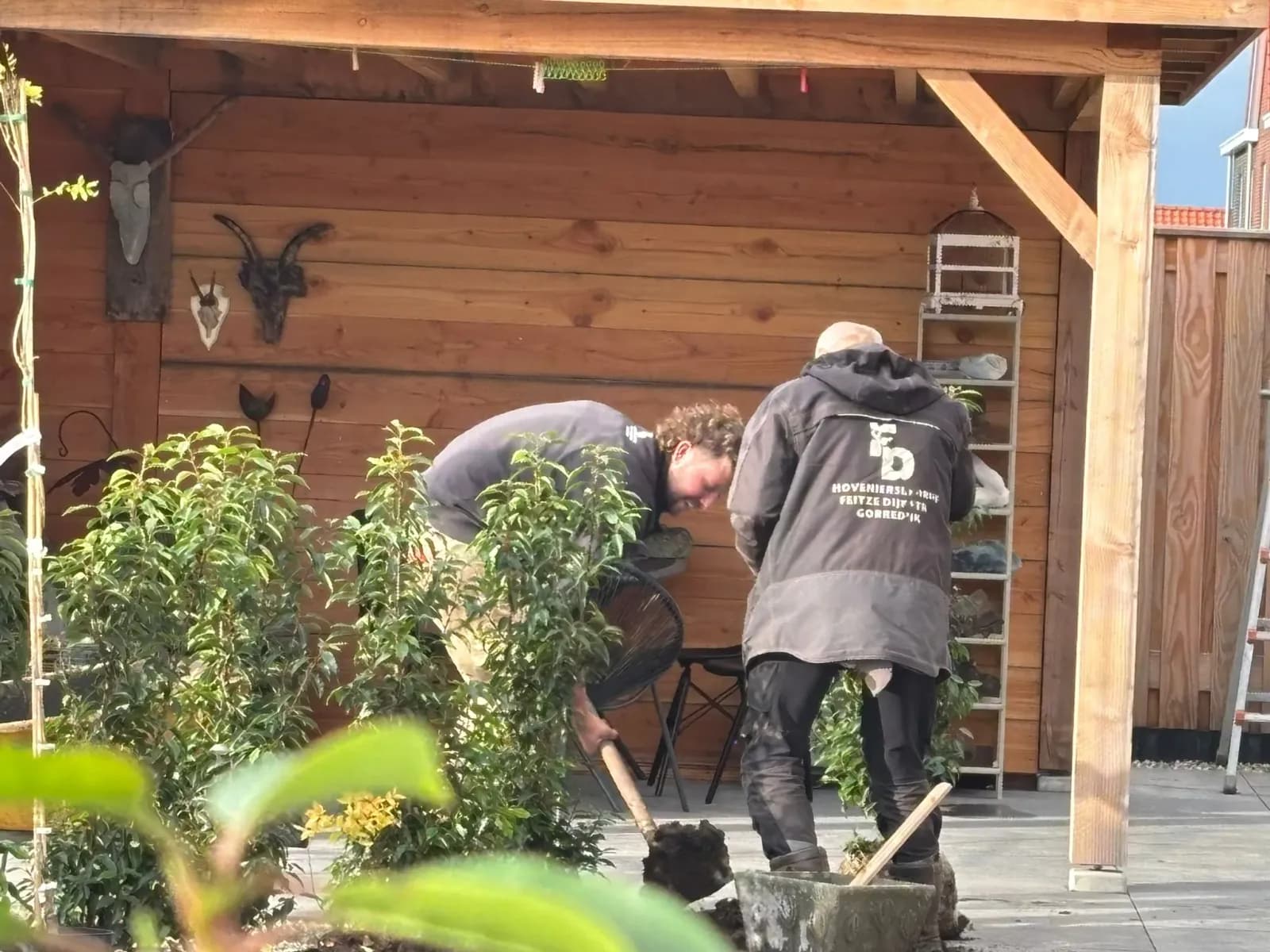 Twee hoveniers in jassen van Hoveniersbedrijf Feitze Dijkstra planten struiken naast de houten terrasoverkapping met terrastegels in De Vrieswijck in Gorredijk