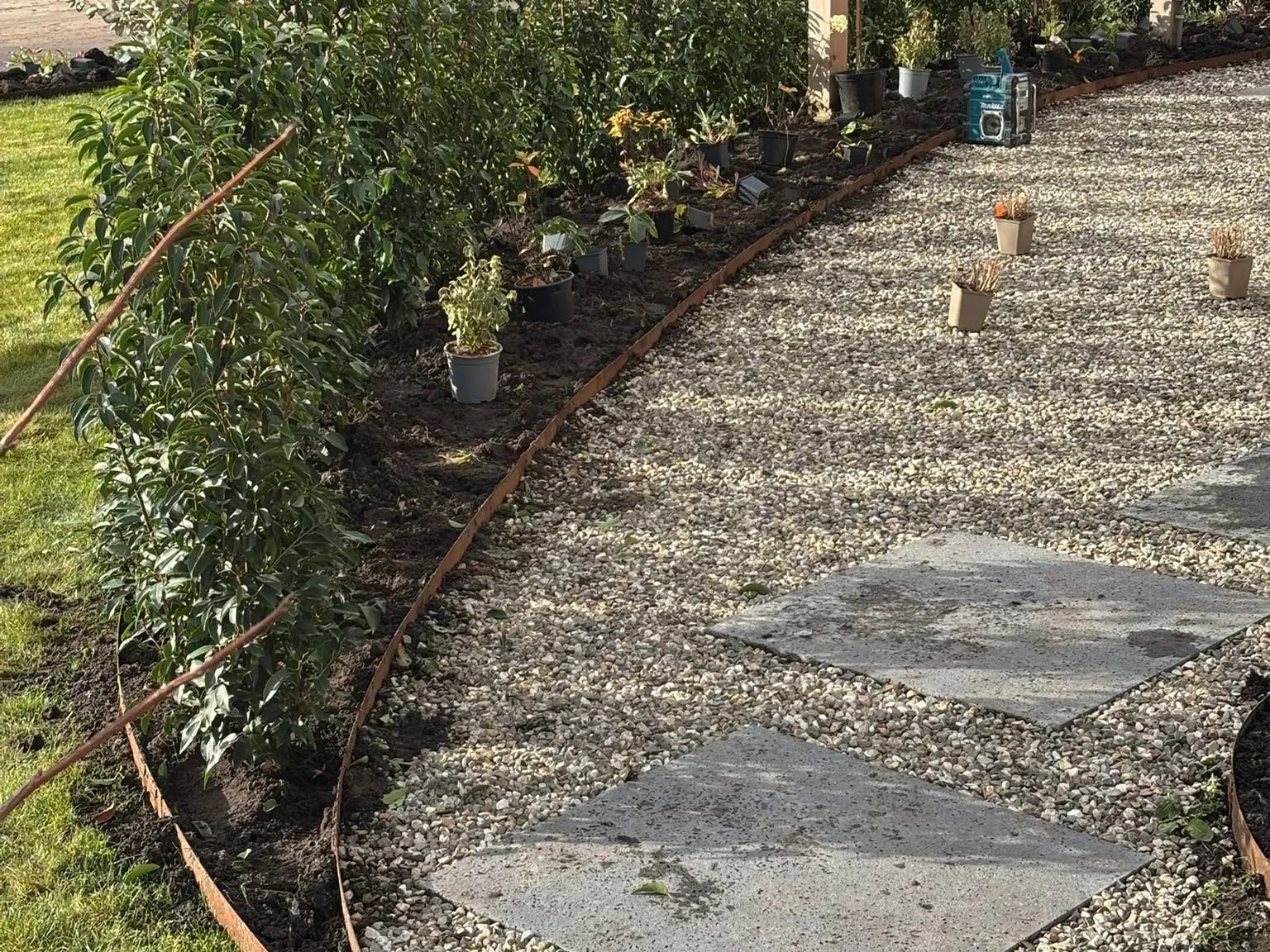 Organisch gevormd grindpad met grote betontegels en een gebogen rand van cortenstaal, jonge struiken langs de rand en een groen gazon ernaast, in een tuin met gras en grind in De Vrieswijck in Gorredijk