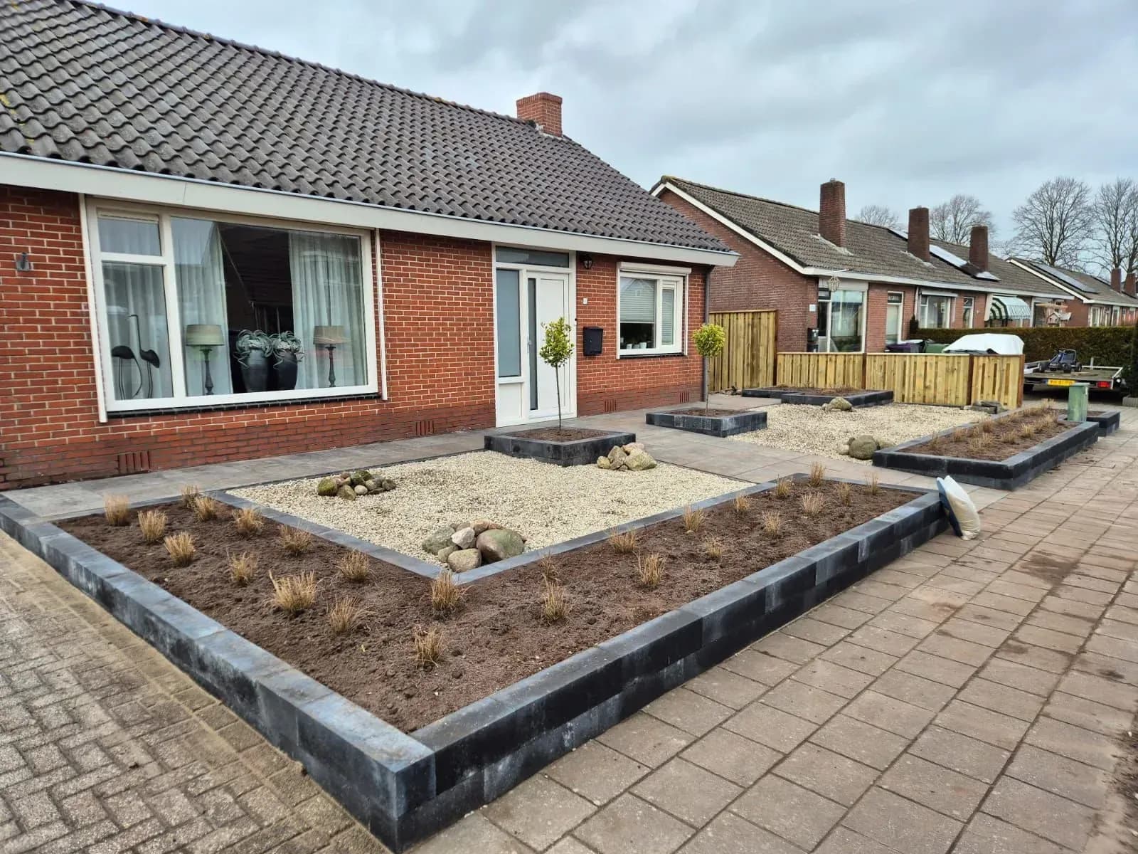 Onderhoudsarme voortuin met grind en verhoogde bakken van stapelblokken, met siergrassen en een bolboompje, bij een vrijstaande woning in Harkema