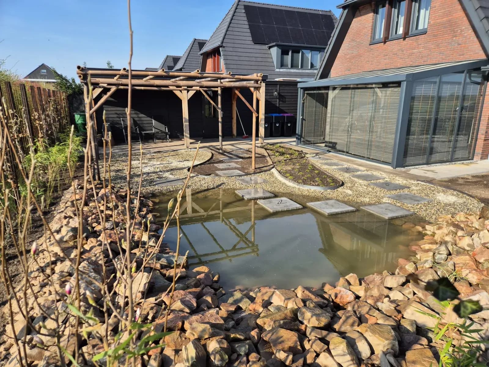 Vijver met rotsen en staptegels, een pergola van bamboe en grind bij een nieuwbouwwoning in Gorredijk, met jonge beplanting eromheen