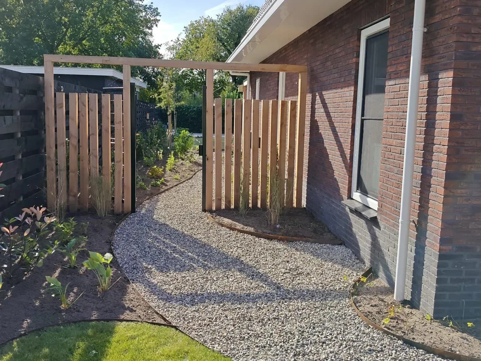Kindvriendelijke nieuwbouwtuin in Lippenhuizen met een gebogen grindpad, houten poort, jonge beplanting en een zwarte schutting