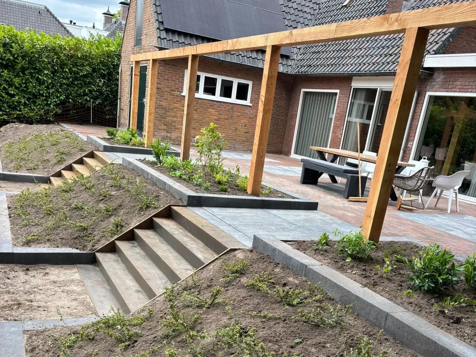 Terras met een tafel en bank onder een pergola van douglas bij een woning in Oldeberkoop, met een trap naar de lagere tuin en jonge beplanting op de taluds