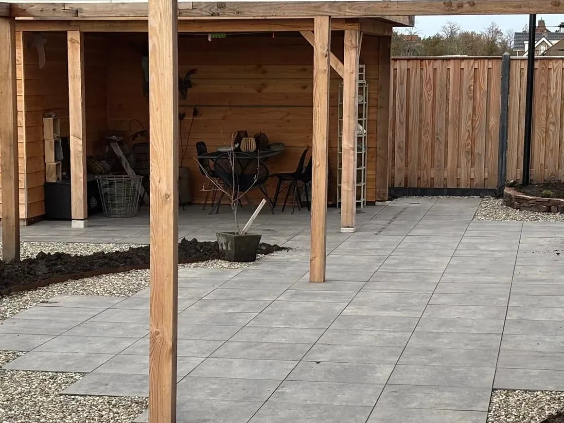 Terras van grijze tegels onder een houten pergola bij een nieuwbouwwoning in De Vrieswijck in Gorredijk, met een overkapping met tafel en stoelen erachter en grind langs het terras. De border links is nog kaal.
