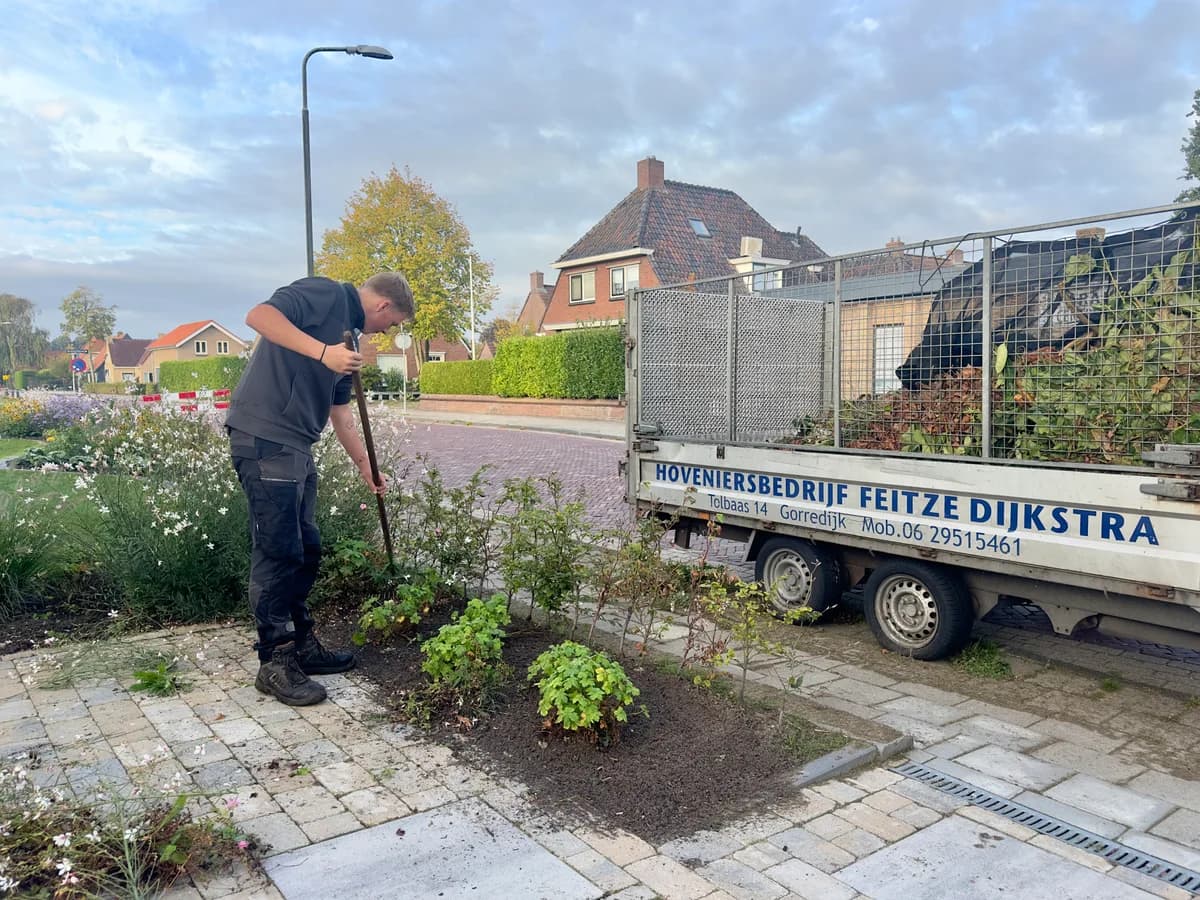 Hovenier werkt in een plantvak langs een oprit van klinkers, naast een aanhanger van Hoveniersbedrijf Feitze Dijkstra vol snoeiafval.