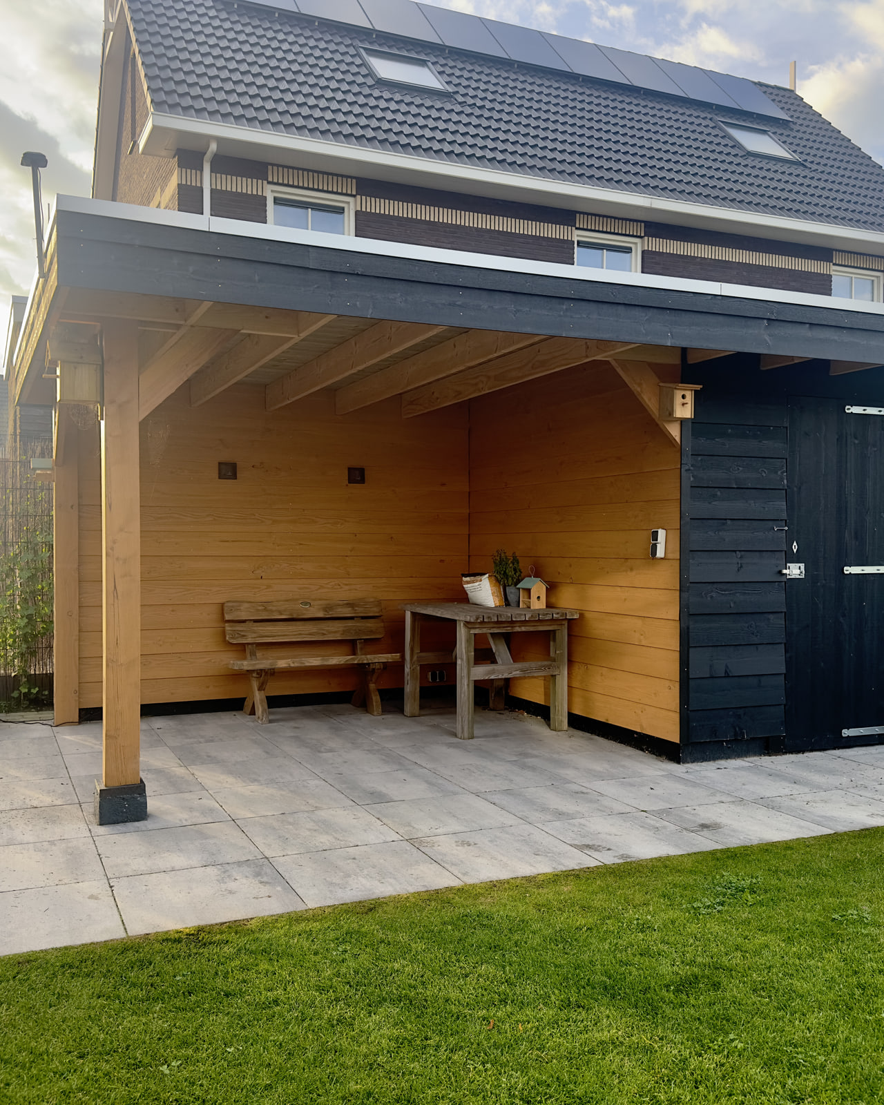Houten overkapping in Gorredijk met een zwarte dakrand en een zwarte berging onder hetzelfde dak, in laag zonlicht. Onder de kap staan een bank en een tafel op een terras van lichte tegels, ervoor ligt een gazon
